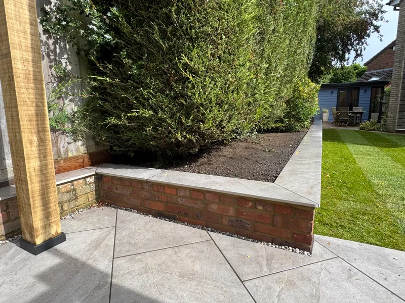 Low red brick raised bed with grey coping stones and a conifer hedge, beside a grey porcelain patio and an oak pergola post