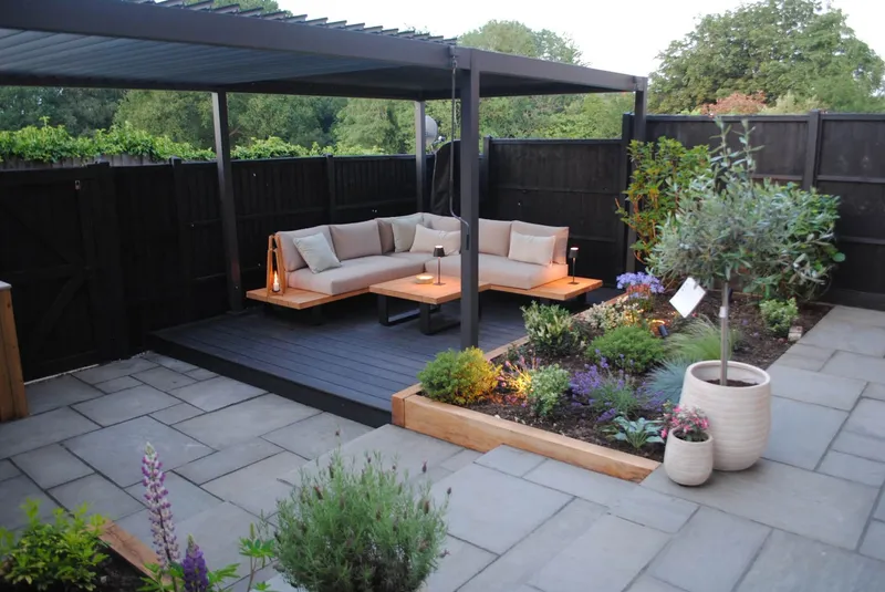 A finished garden design and build, with grey paving, a raised timber planter and a dark deck with outdoor sofas under a louvred aluminium pergola