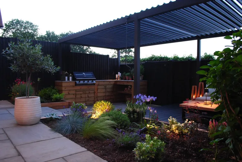 The finished garden at dusk, with lit planting and a timber-clad outdoor kitchen with a built-in barbecue beneath the pergola